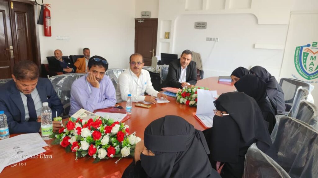 Group of people in formal attire seated around a conference table with red and white flower arrangements, reviewing documents in a meeting room.