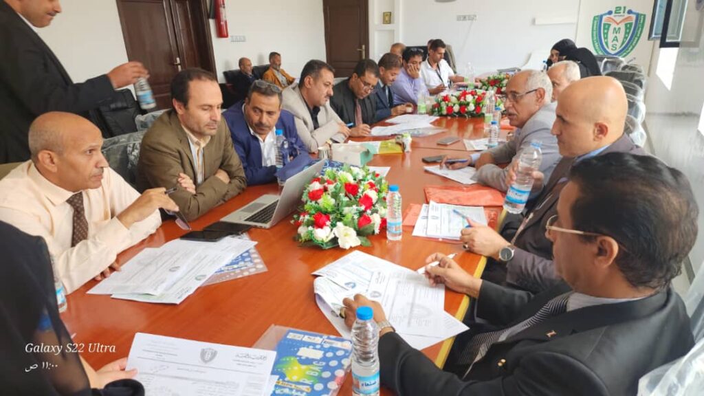 Group of professionals in a boardroom around a long conference table, reviewing documents, laptops, water bottles, and a floral centerpiece.
