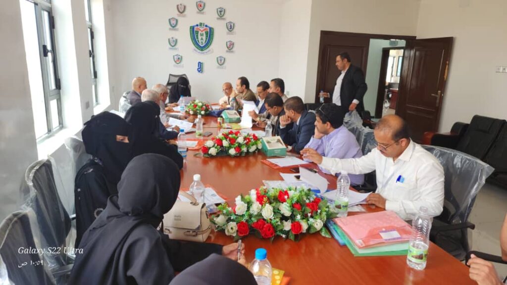 Group of professionals around a long conference table in a bright room, with floral centerpieces and emblems on the wall.