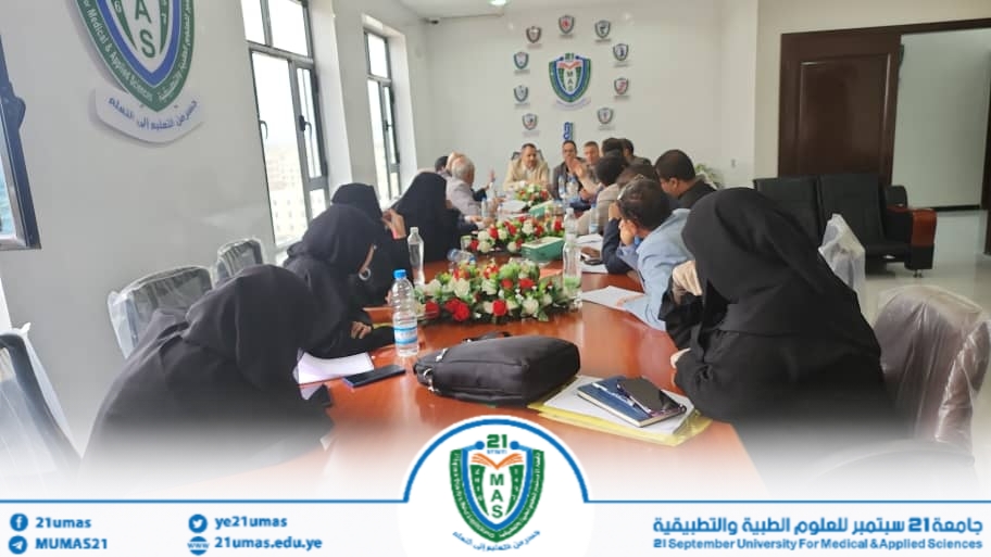 People seated around a long conference table in a university meeting room, with a floral centerpiece and a crest on the wall, engaged in discussion.
