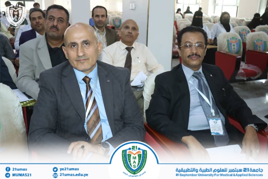 Two men in suits sit in the front row of a university conference, with others behind them and event branding visible at the bottom banner.