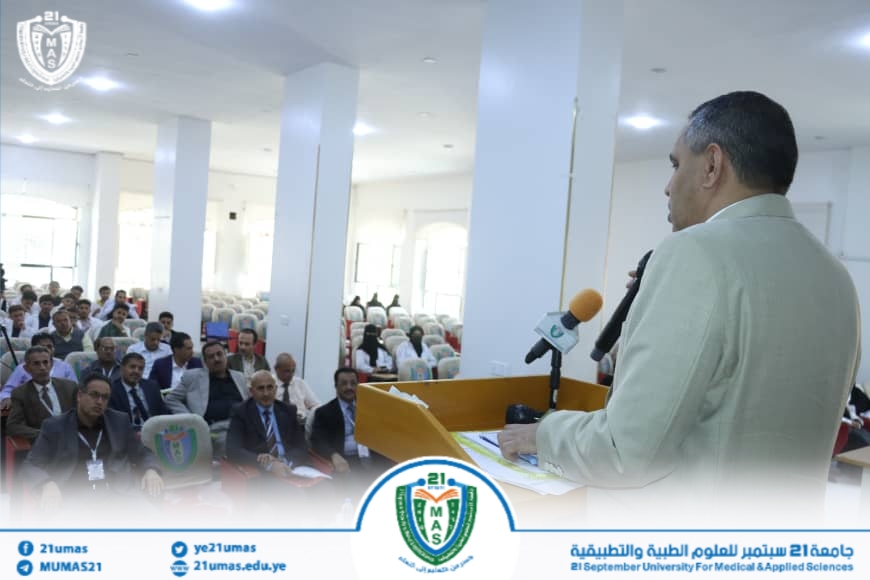 Speaker at a podium addressing a seated audience in a bright conference hall, university banner and logos visible at the front.