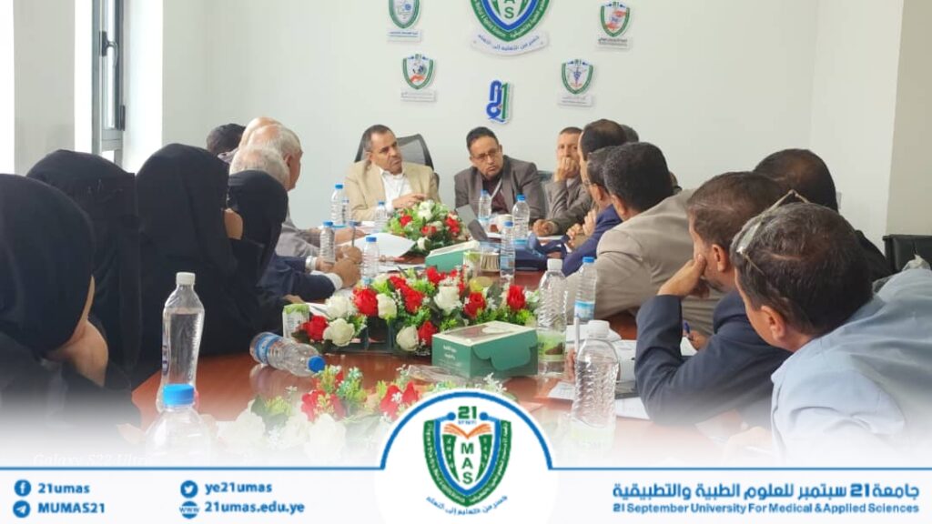 Group of professionals seated around a long conference table in a formal meeting, with a floral centerpiece and water bottles, banners and university logos on the wall behind them.
