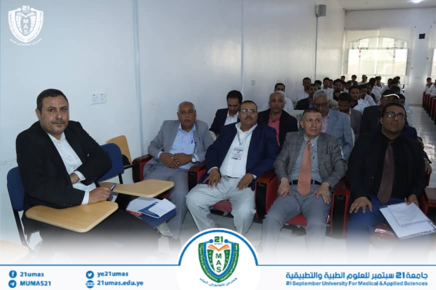 Group of men seated in a classroom auditorium, facing forward, during a formal meeting at 21 September University for Medical & Applied Sciences. A banner with the university logo is at the bottom.