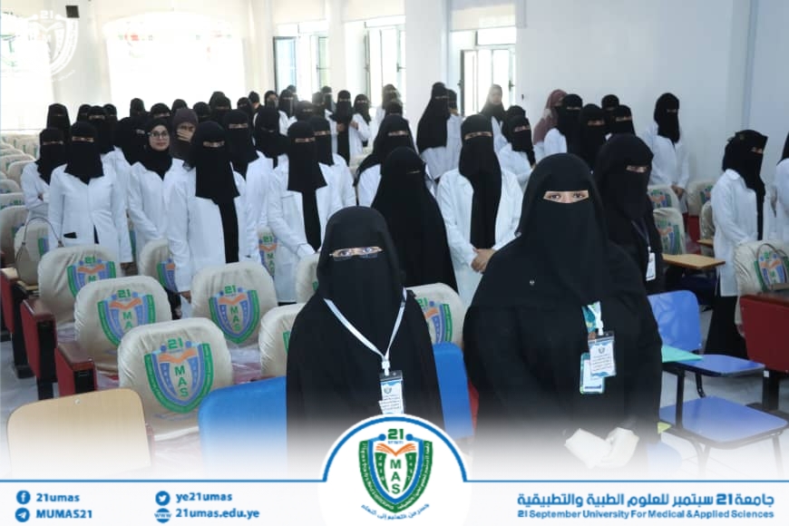 Group of hijab-wearing female students in white coats standing in a university hall during an event, with badges visible.