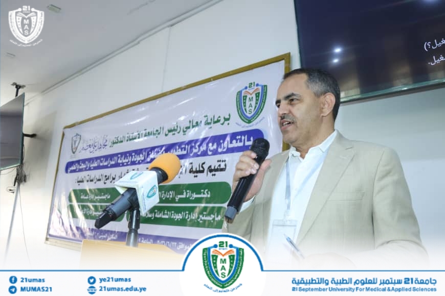 Man in a beige suit giving a speech at a podium during a university conference, with microphones and a banner with Arabic text and logos in the background.