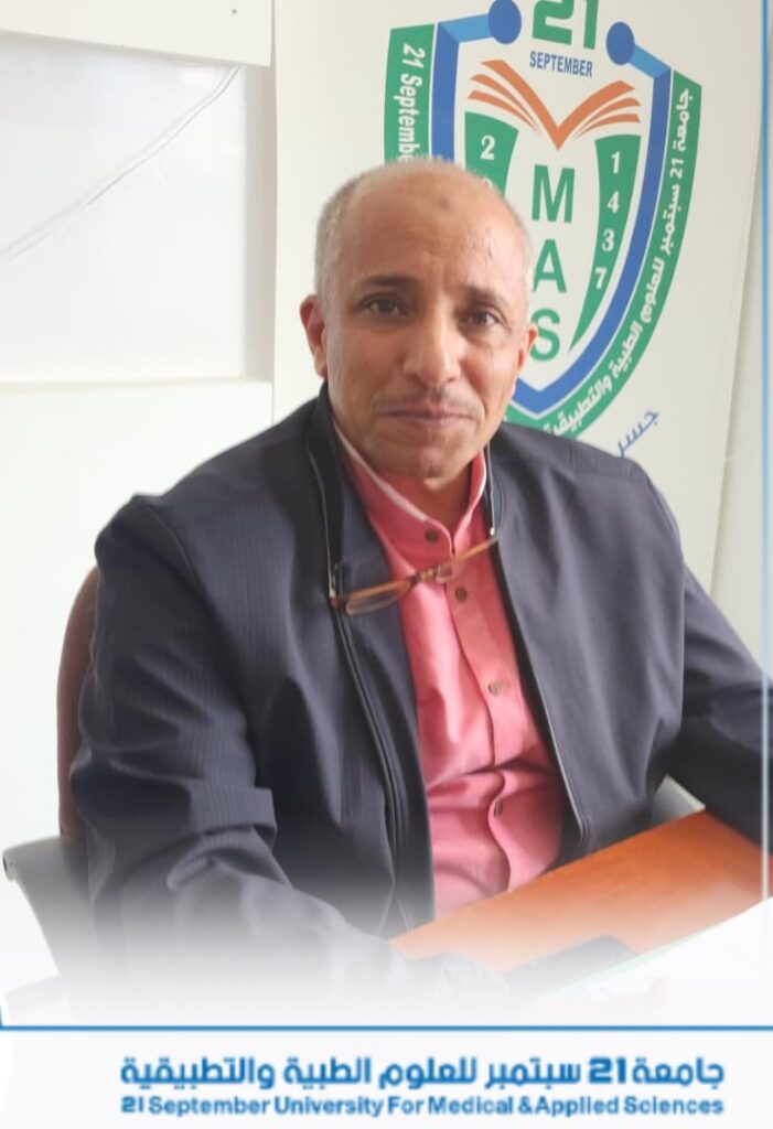 Portrait of a man in a pink shirt and dark jacket seated at a desk, with a banner in the background that reads'21 September University For Medical & Applied Sciences'