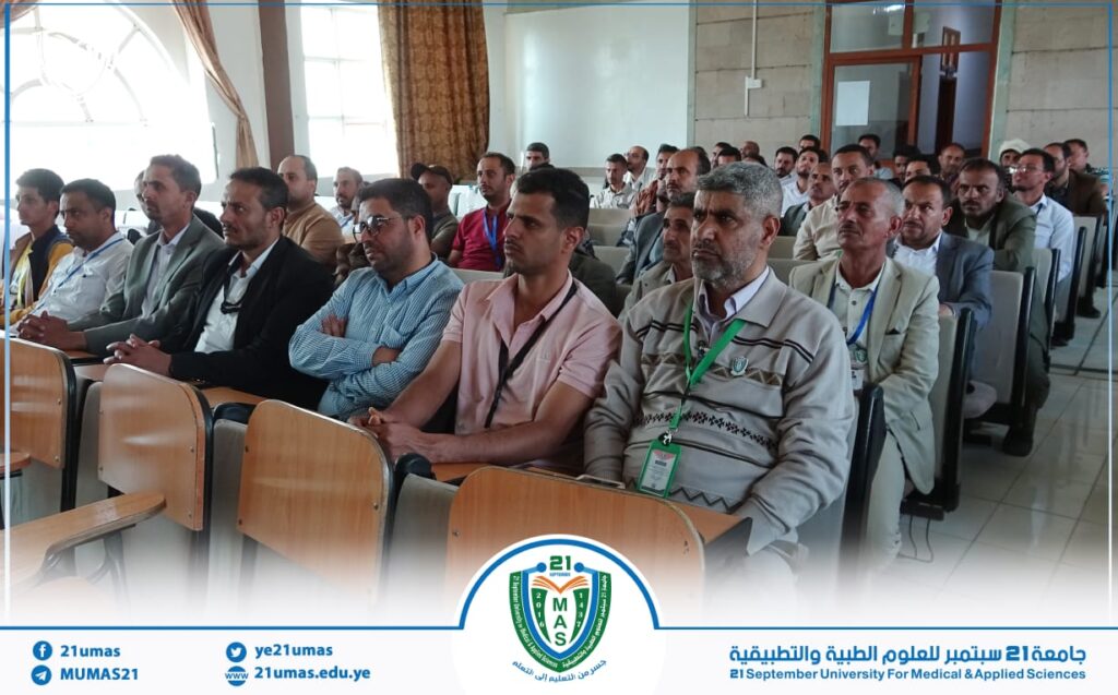 Group of men seated in a lecture hall listening to a presentation, wearing conference badges; university logo and banner visible at the front.