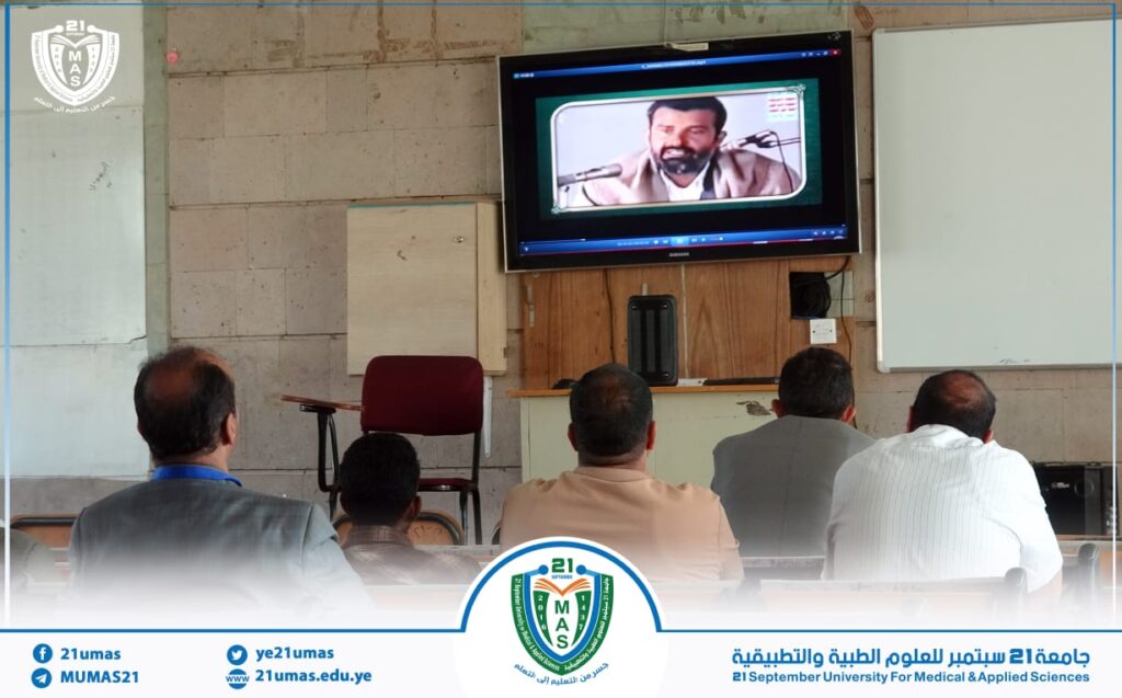 Students seated in a classroom watching a video lecture on a wall-mounted monitor.