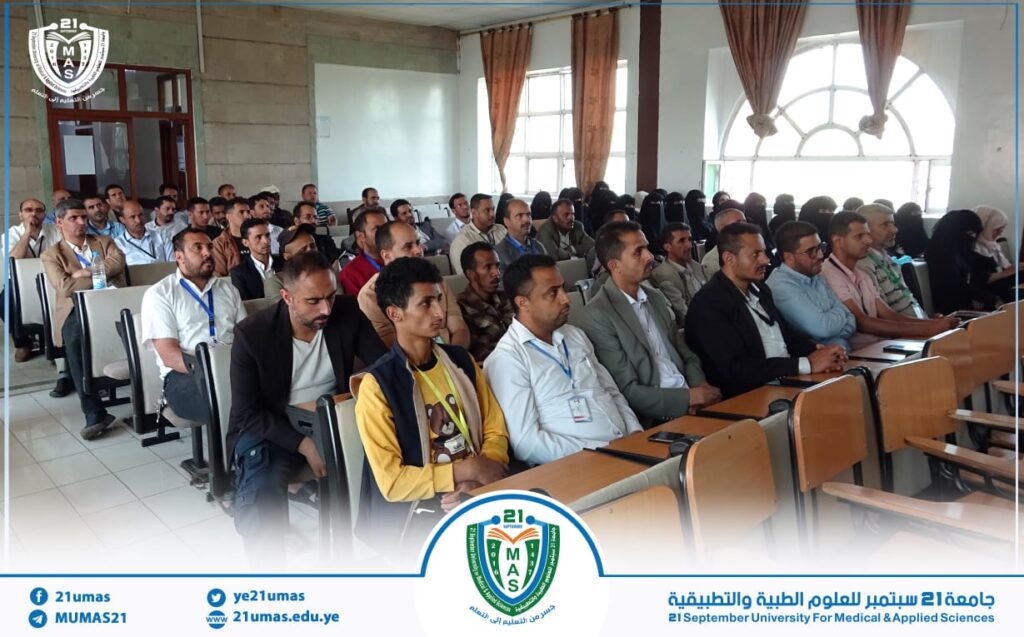 Audience of men and women seated in a classroom-style lecture hall during a university event, with the university crest visible on the bottom banner.