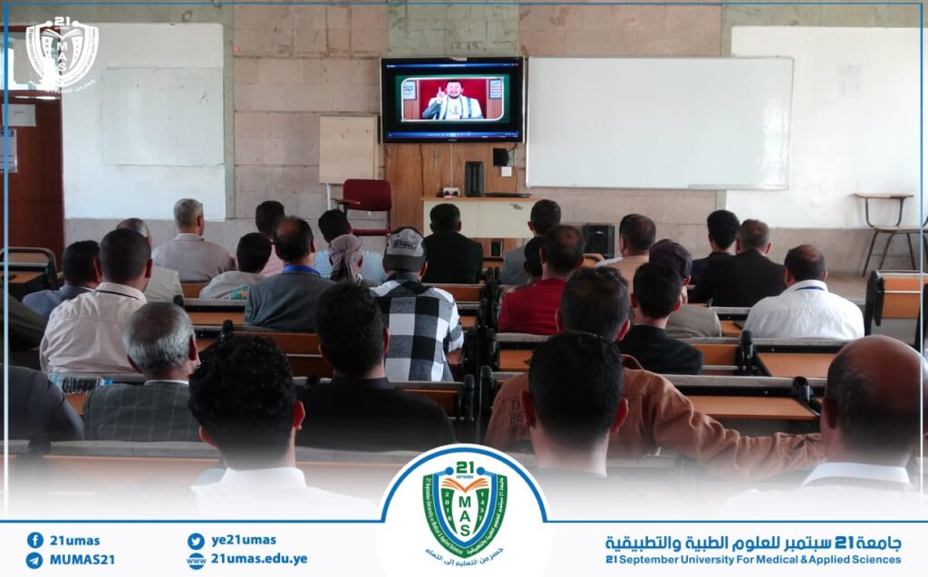 Classroom audience watching a video lecture on a wall-mounted TV screen at the front of a lecture hall. Whiteboard behind and university branding at the bottom border.