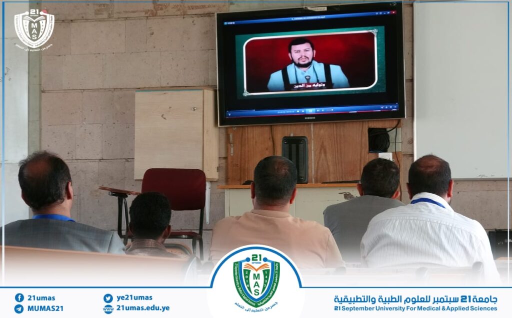 People seated in a classroom watching a video of a man on a wall-mounted screen; university branding on the bottom border indicates a session at 21 September University for Medical and Applied Sciences.