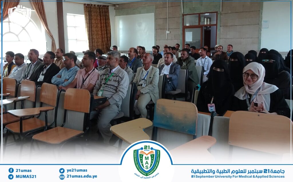 Audience seated in a lecture hall during a university event, with many men and women in traditional clothing and hijabs, banner visible at bottom.