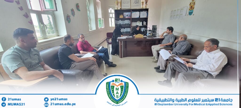 Group of people seated in a meeting room around a desk, some holding documents, with two women in hijabs at the desk, in a university office setting. A university banner with the 21 September University for Medical & Applied Sciences logo is visible at the bottom.