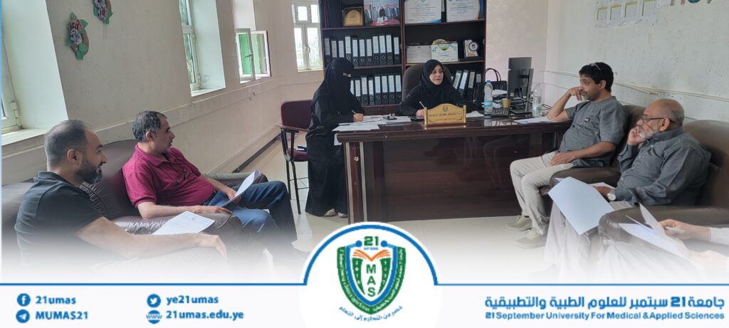 Six people in a meeting in an office: two women in hijabs behind the desk and four men seated with papers around the room, university branding visible at bottom edge.