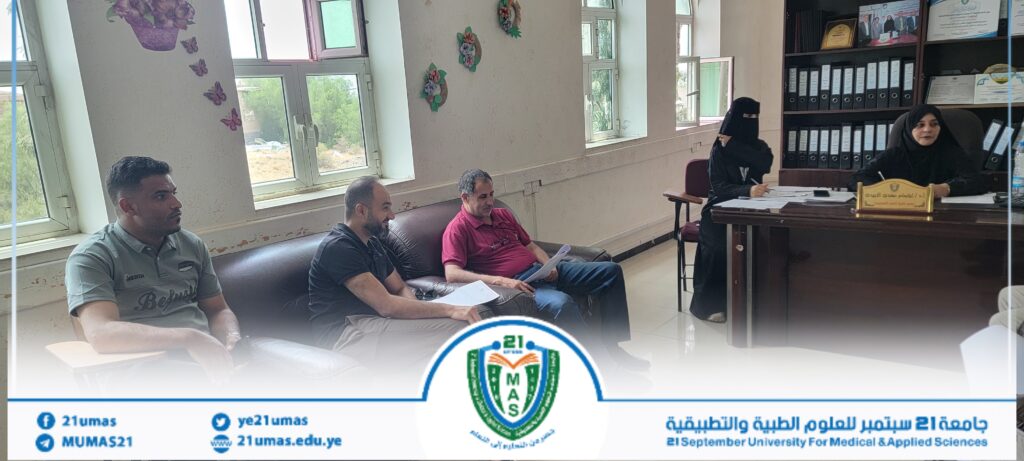 Three men sit on a sofa in a meeting room while two women in hijabs work at a desk to the right, with a university banner and emblem visible at the bottom.