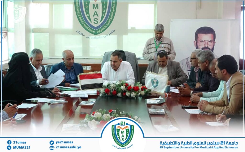 Group of officials seated around a conference table during a formal meeting at 21mas university, with MAS banner and a portrait in the background