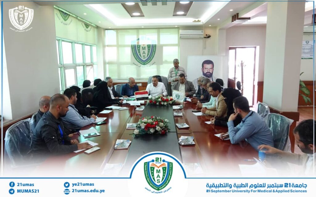 Group of professionals seated around a long conference table in a formal university meeting room, MAS emblem on the wall behind them.