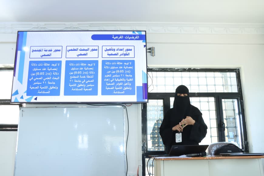 Female presenter wearing a niqab giving an Arabic-language lecture in a classroom, with a blue Arabic slide projected on a screen behind her.