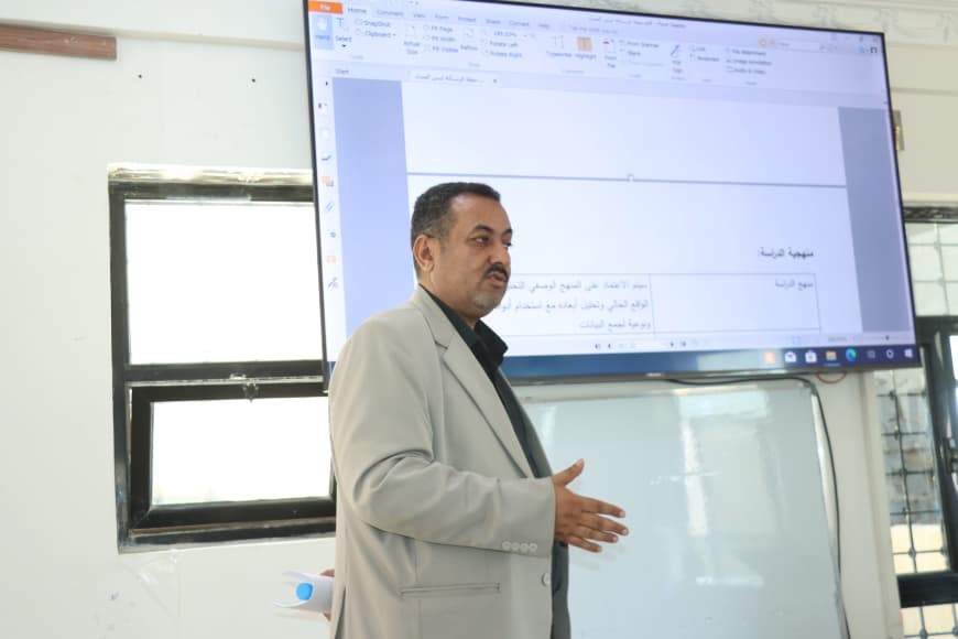 A man in a light gray blazer presents in a classroom, gesturing with his hand as slides are projected on a screen behind him.