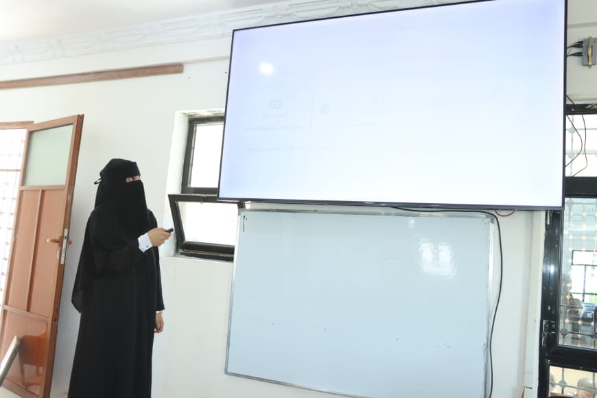 Woman in a black abaya/niqab points at a large wall-mounted screen in a classroom.