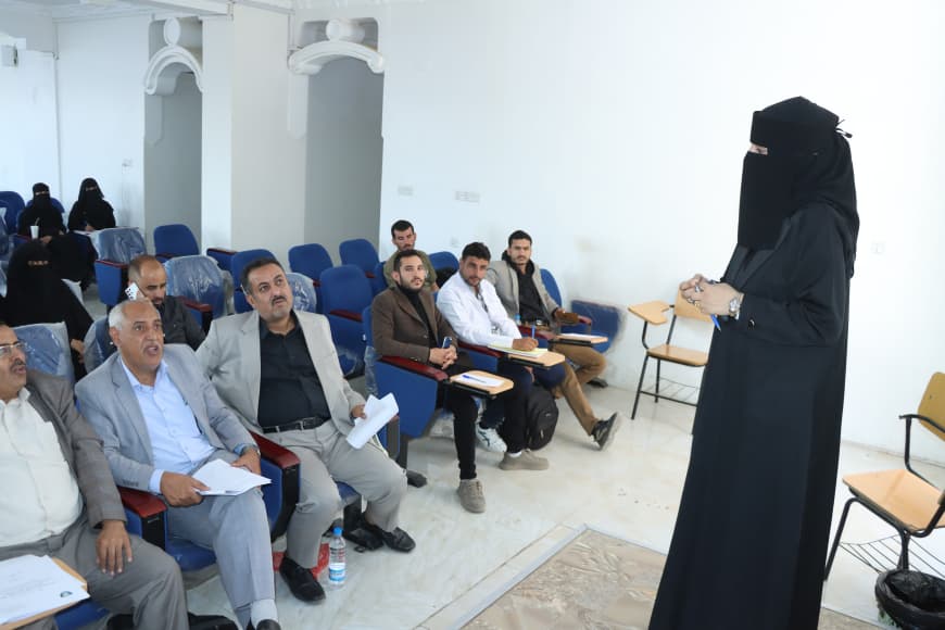 A woman wearing a black niqab stands at the front of a classroom addressing a seated group of men with notebooks.