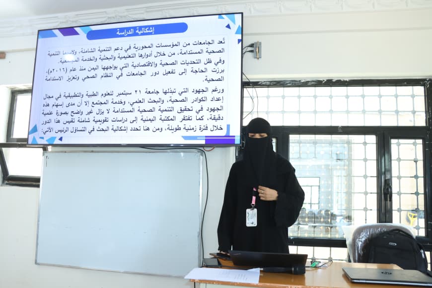 Woman in a black niqab stands at a desk in a classroom, presenting a large Arabic slide and a whiteboard behind her.