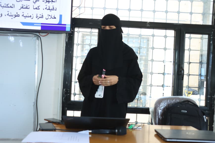 Woman in a black abaya and niqab standing behind a desk, holding a small bottle in an office setting with a laptop and papers on the desk.