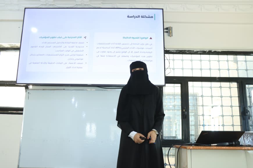 A woman in a black abaya and niqab stands at the front of a classroom, presenting beside a whiteboard with an Arabic slide projected on a screen behind her.
