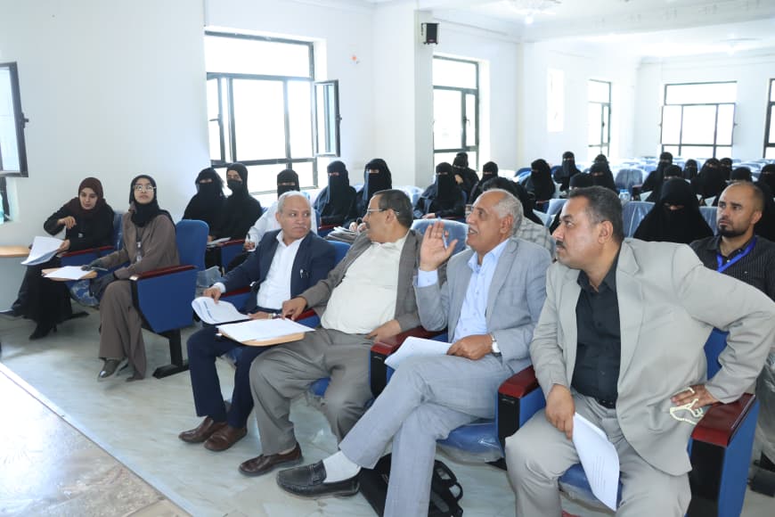 Front row of attendees in a seminar: men in suits with papers, while women in hijabs sit in the background.