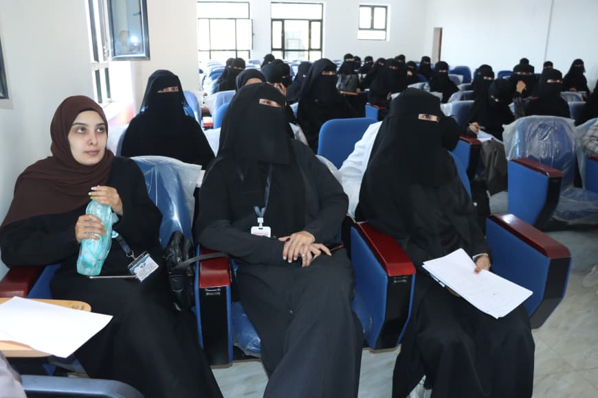 Group of women wearing black abayas and niqabs seated in a lecture hall; one foreground woman holds a water bottle as she looks to the side.