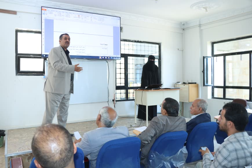 Lecturer in a light gray suit speaks to an audience in a classroom, a large screen showing a presentation behind him while attendees sit in blue chairs.
