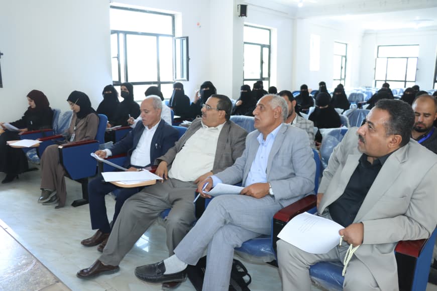 Audience in a bright classroom: front row of men in suits with papers, women in black hijabs sit behind them.