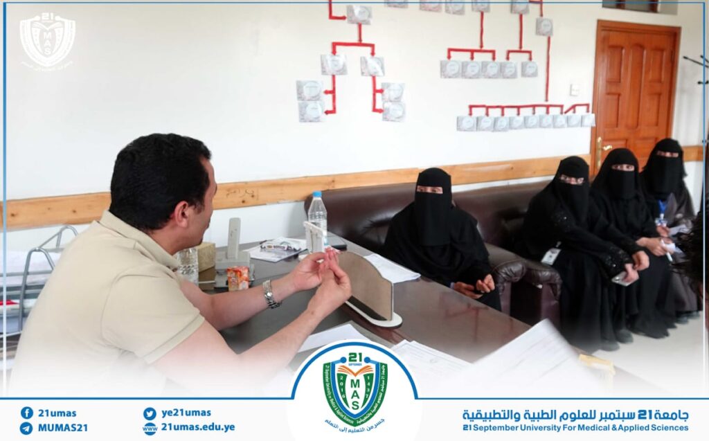 Man in a beige shirt sits at a desk explaining something to women wearing niqabs in a classroom/office setting, with charts on the wall behind them.
