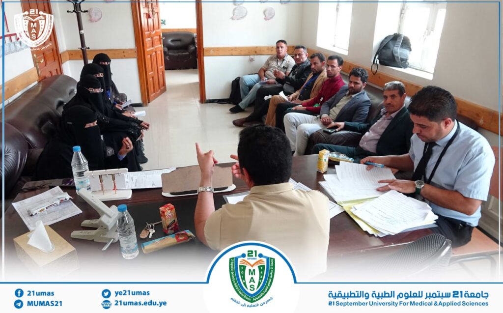 Group of men in suits and women in black abayas sit around a conference table in a university office, discussing documents and notes.
