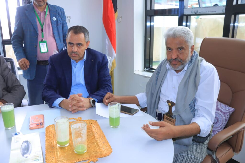 1001114297 - جامعة 21 سبتمبر Three men sit around a table with green drinks; one man in a blue suit and another elderly man with a scarf and cane talk, while a standing man watches in a blue jacket near a flag in the background.