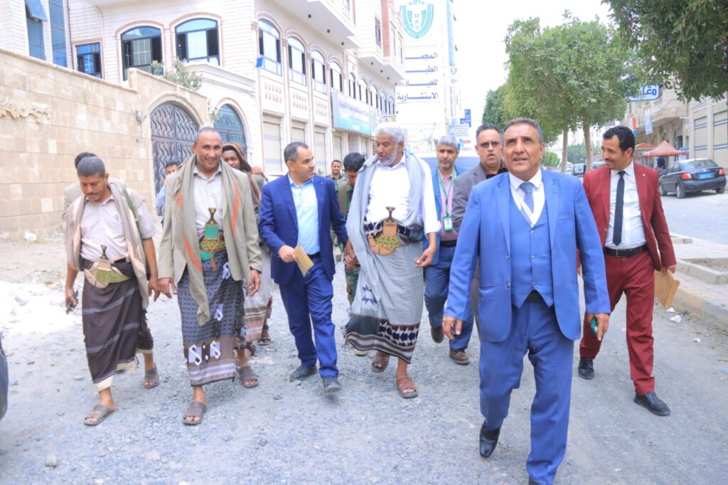 1001114300 - جامعة 21 سبتمبر Group of men walking together down a uneven street, some in traditional robes and sandals and a few in suits, as they chat and stroll the sidewalk.