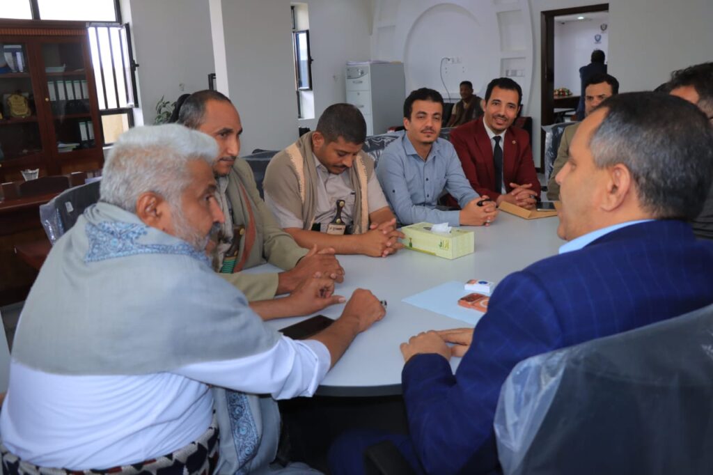 1001114303 - جامعة 21 سبتمبر Group of men seated around a conference table in a meeting room, discussing and leaning forward in conversation.