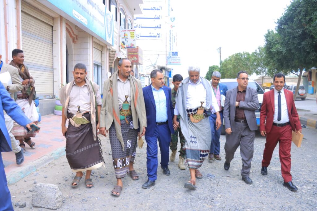 1001114309 - جامعة 21 سبتمبر Group of men walking down a street; several wear traditional Yemeni attire with jambiya daggers, others in suits, in a market area.