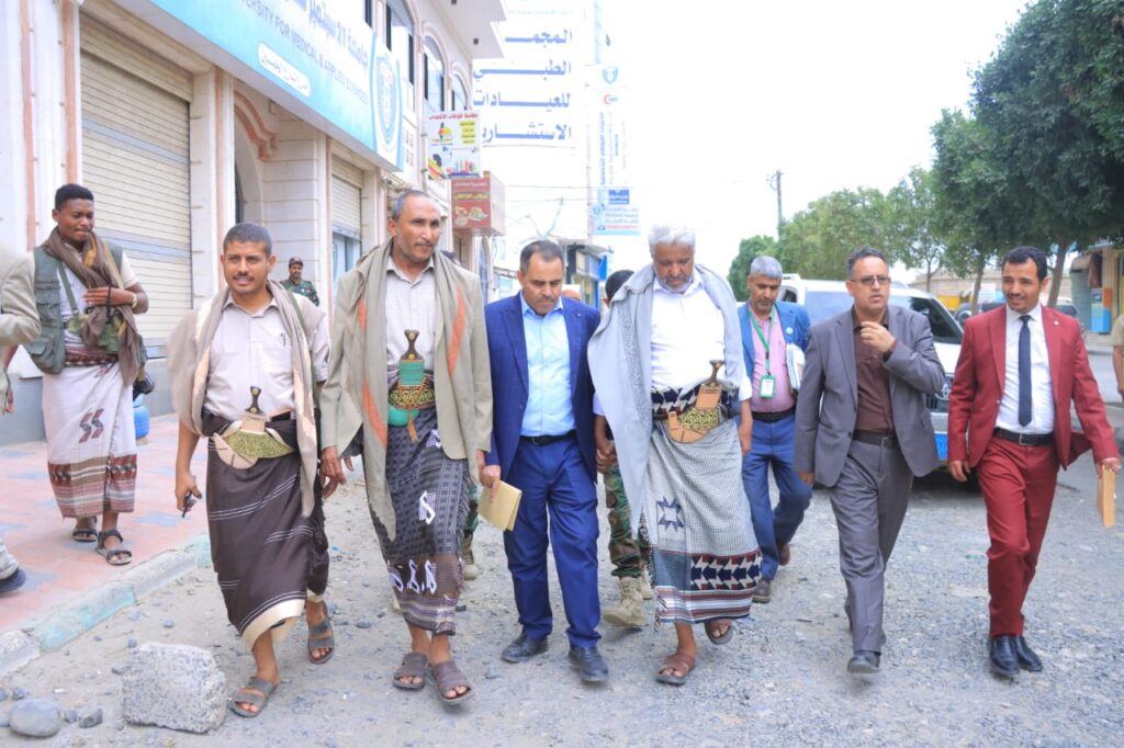 1001114318 - جامعة 21 سبتمبر Group of men walking down a rough street; several wear traditional robes with curved dagger sheaths at their waists.