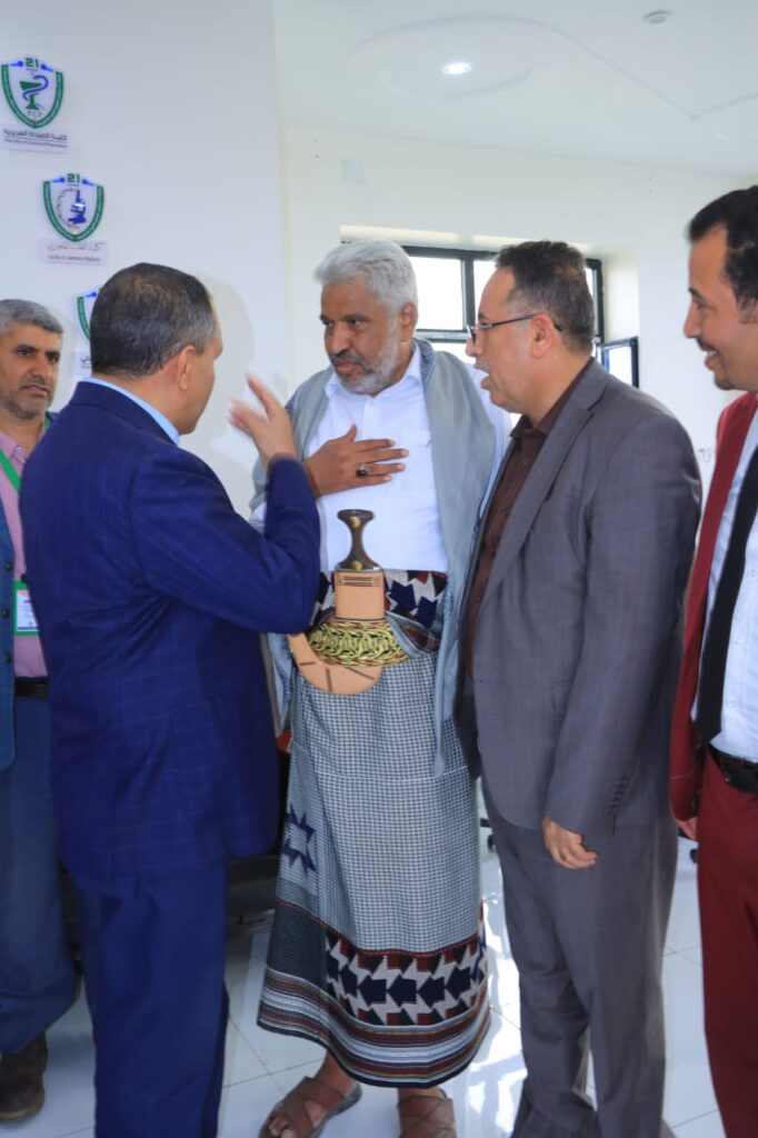 1001114321 - جامعة 21 سبتمبر Five men in business attire talk in a bright room; a man in a blue suit gestures while an older man in traditional robe holds a decorative trophy vase.