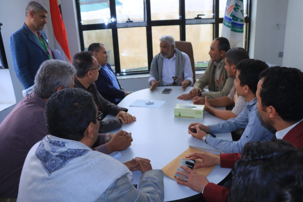1001114327 - جامعة 21 سبتمبر A group of men sit around a white oval conference table in a bright room, one man at the head speaking with hand gestures while others listen; flags are visible near the windows.