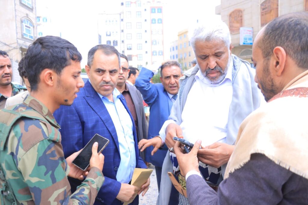 1001114330 - جامعة 21 سبتمبر Group of men in a city street examining a small handheld device or phone together, some holding documents, uniforms and suits suggest a briefing or inspection.