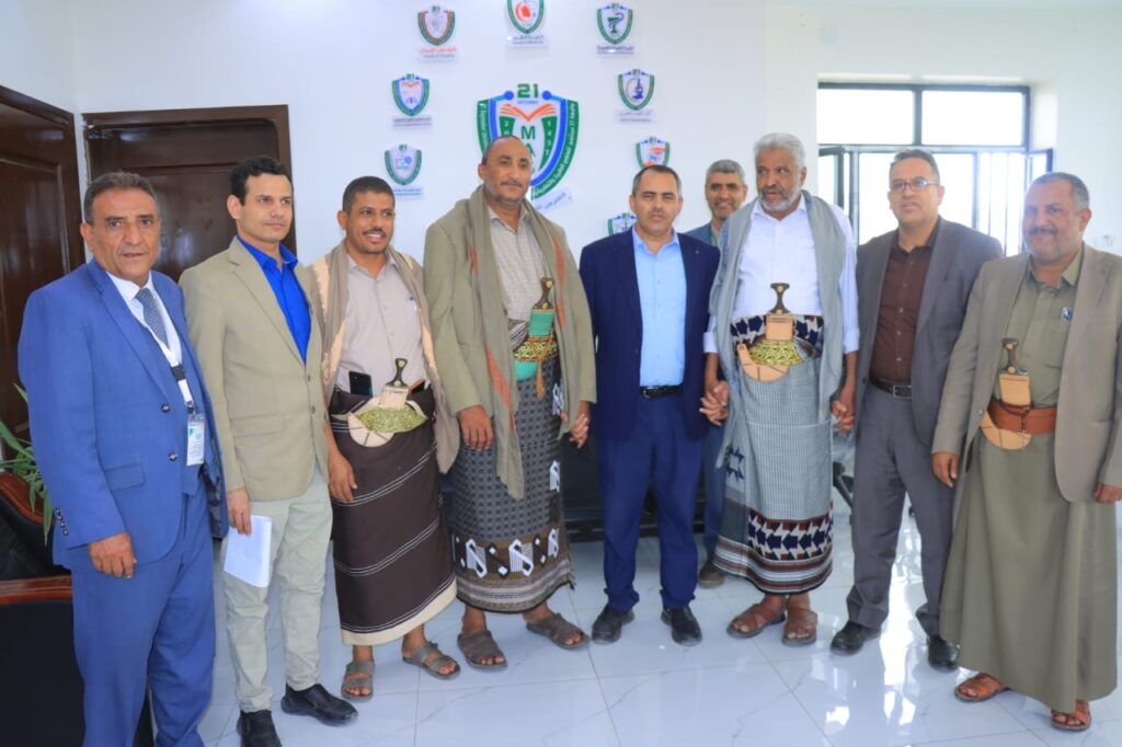 1001114336 - جامعة 21 سبتمبر Group of nine men posing for a formal photo indoors, some in traditional attire with ceremonial daggers, logos on the wall behind them.