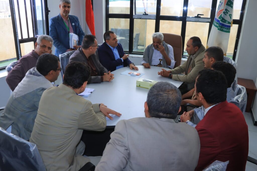 1001114339 - جامعة 21 سبتمبر Group of men in suits sits around a large conference table in a bright office, having a discussion and listening to one speaker.