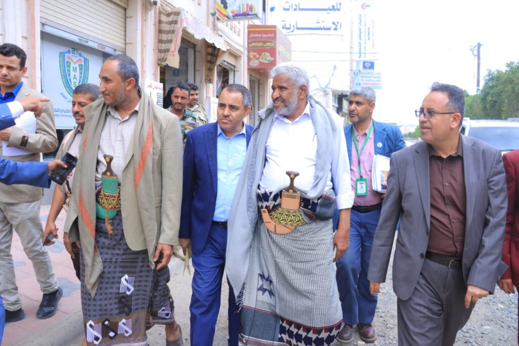 1001114347 - جامعة 21 سبتمبر Group of men in traditional clothing and suits walking together on a street during a visit or outdoor event. A man in front wears a scarf and a sword sheath as part of traditional attire.