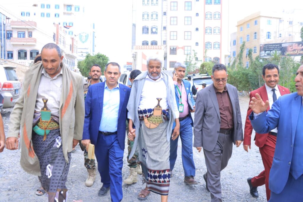 1001114364 - جامعة 21 سبتمبر Group of men in suits and traditional robes walking together on a gravel urban street, with two men wearing ceremonial daggers at their waists.