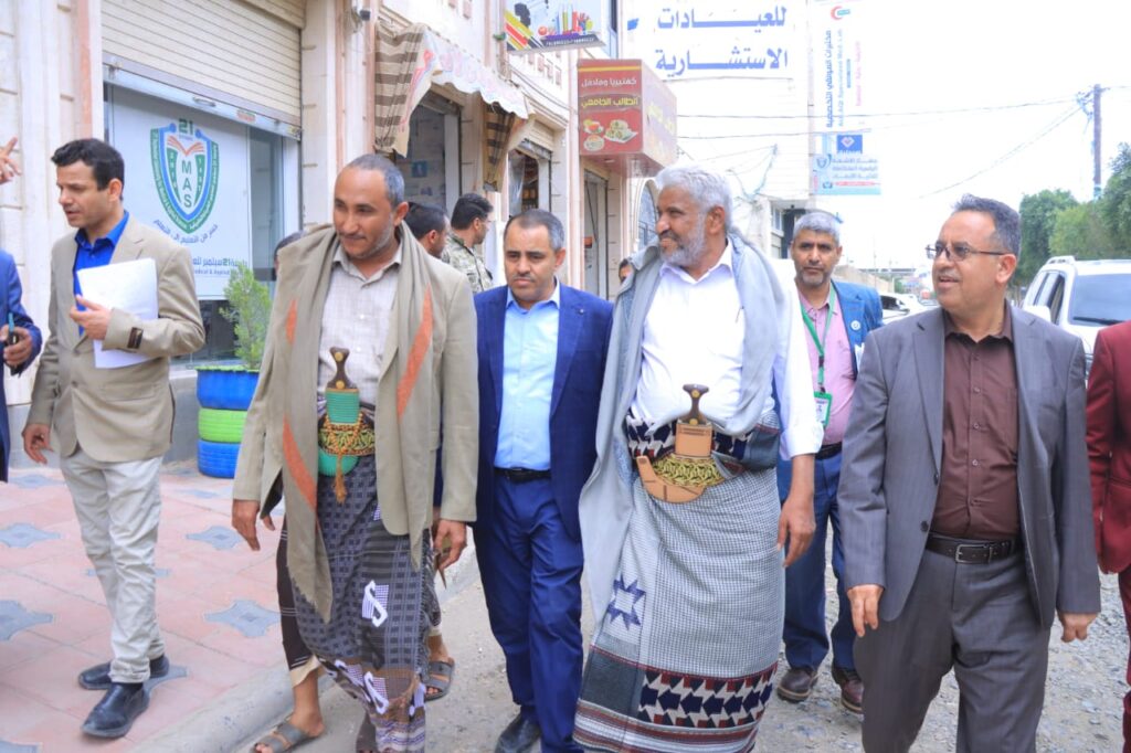 1001114375 - جامعة 21 سبتمبر Group of men walking down a storefront street; two wear traditional robes with ceremonial daggers at their waists.