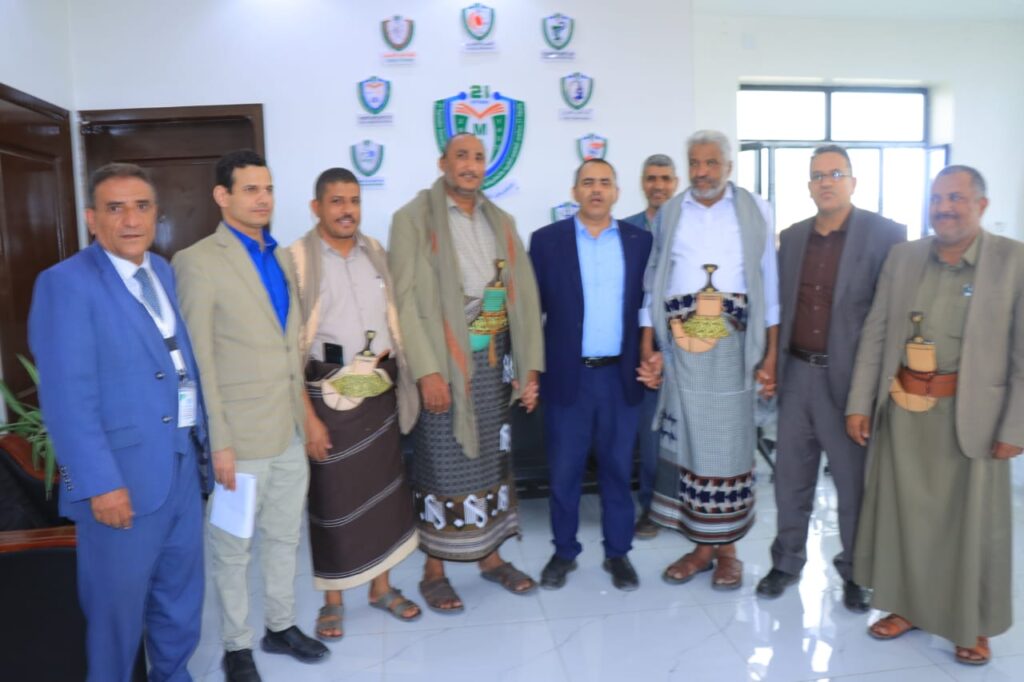 1001114384 - جامعة 21 سبتمبر Group of men posing for a photo indoors, several wearing traditional Yemeni clothing and others in suits, against a wall with logos.