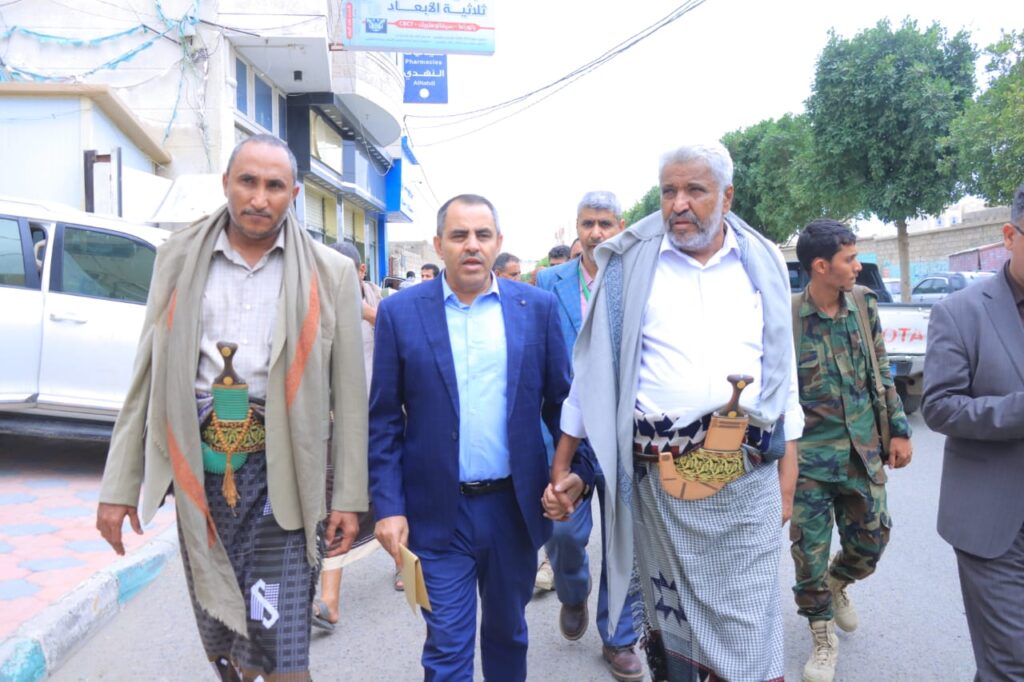 1001114390 - جامعة 21 سبتمبر Group of men walking on a city street; two older men wear traditional robes with daggers at the waist, flanked by others in suits.